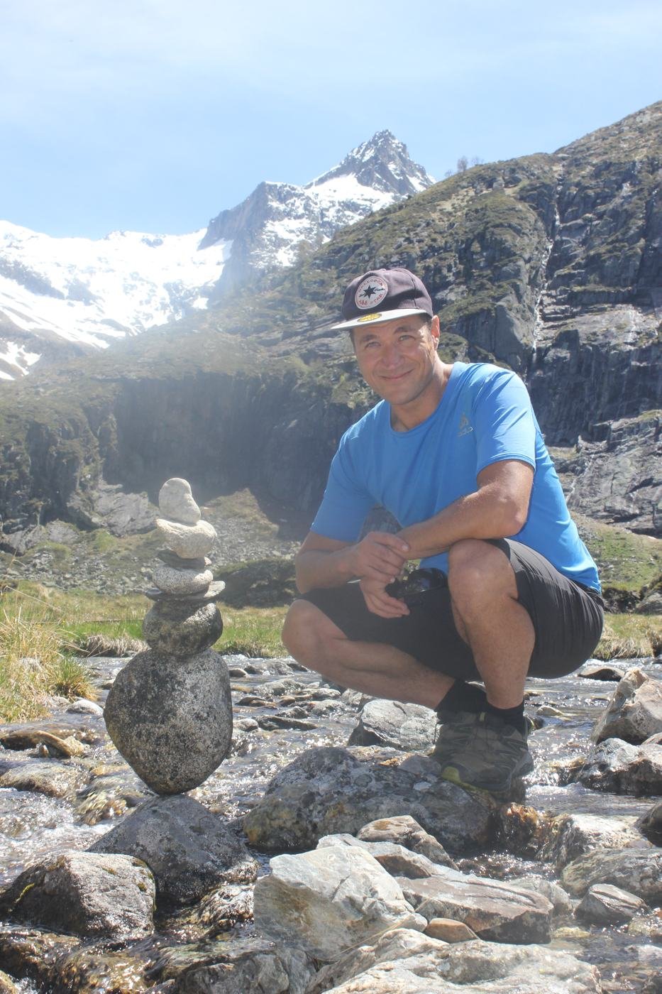 Christophe accroupi près d'un cairn au bord d'un torrent, sommets enneigés derrière